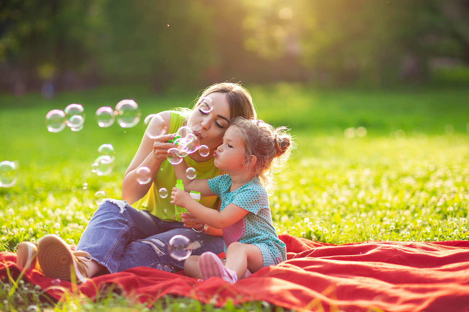A smiling mother and her young daughter blowing bubbles together on a sunny day in a London park.