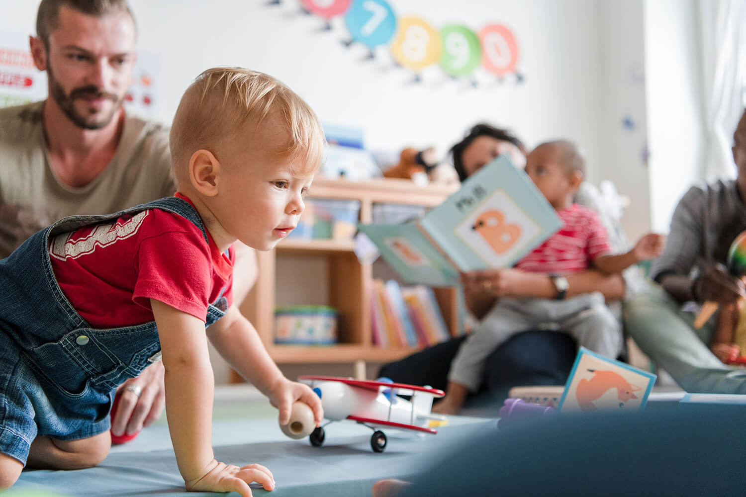 A toddler plays with a wooden toy plane in a family-friendly learning space while parents read to other children.