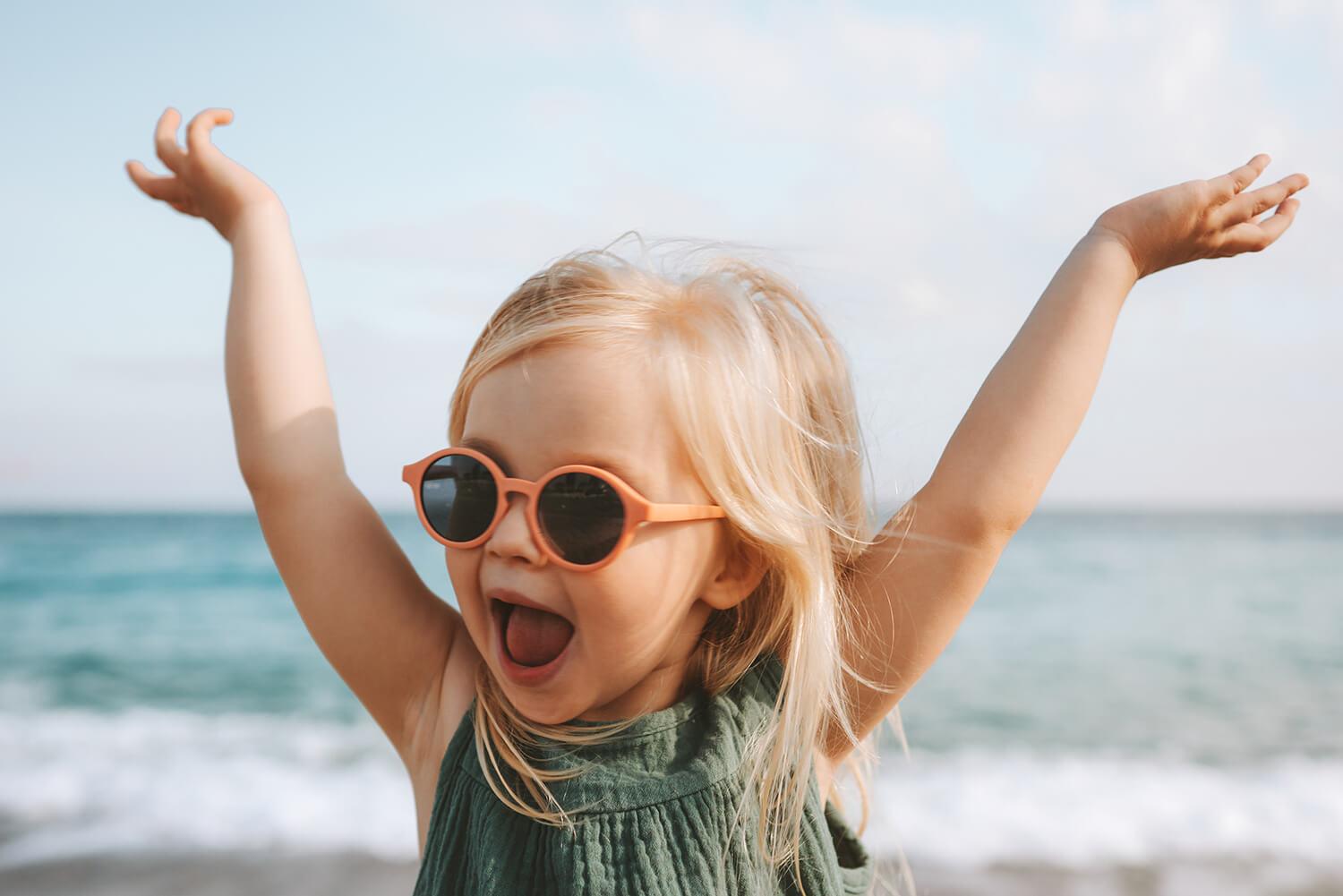 Joyful blonde girl in sunglasses raising her arms while standing on a beach during a sunny holiday near London.